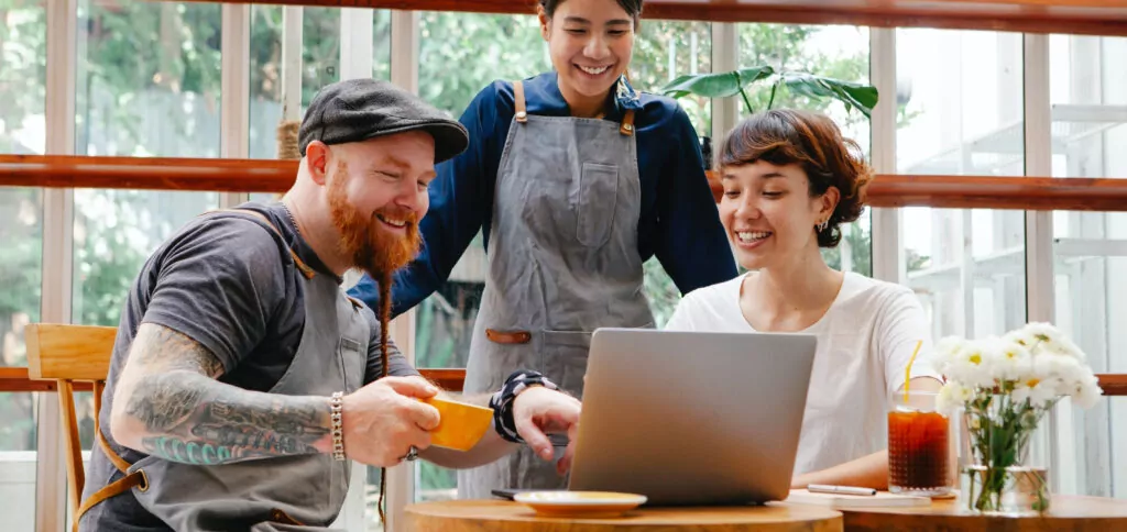 Three people gathered around a laptop in a bright cafe setting, collaborating and sharing ideas. Natural light streams through windows, creating a warm, creative atmosphere. Fresh drinks and flowers on the wooden table add to the casual, productive environment.