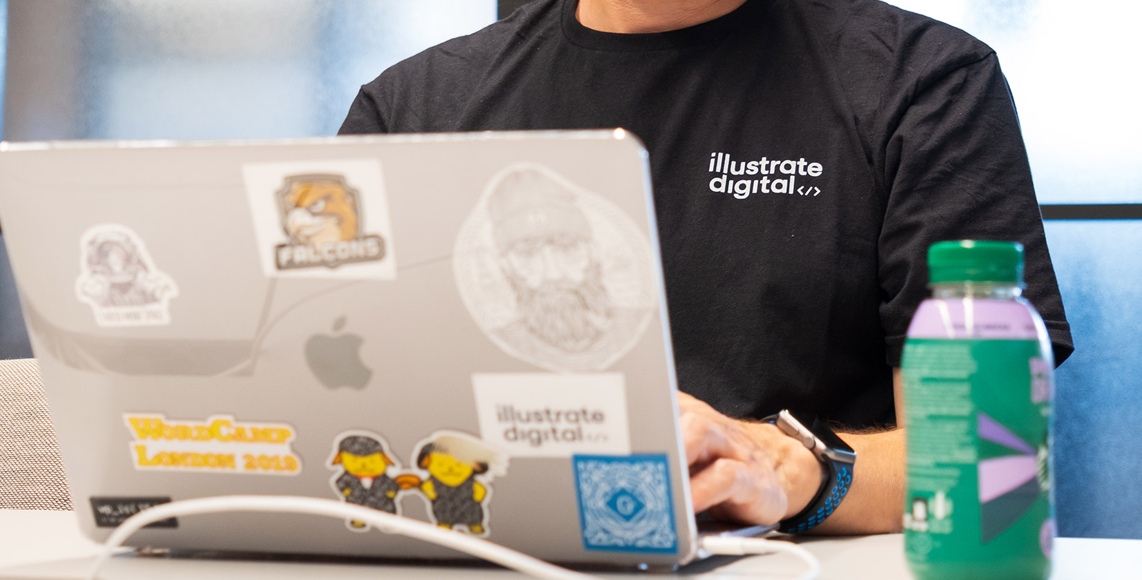 A picture of a developer at Illustrate Digital coding on their laptop, the laptop has various industry stickers on the back including the Illustrate Digital logo